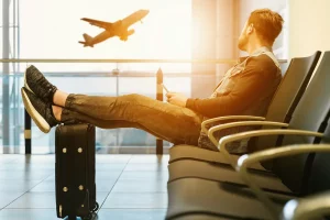 A man sitting in an airport lounge with his feet up on his suitcase looking out of the window looking at a plane taking off
