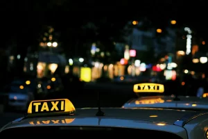 The roofs of 2 taxis at night with their lights illuminated on top and a street view behind