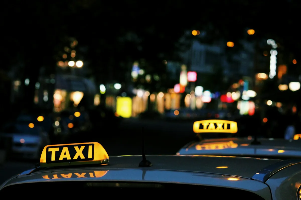 The roofs of 2 taxis at night with their lights illuminated on top and a street view behind